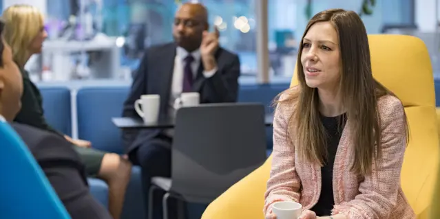 Woman in pink jacket seated in yellow chair in modern office with colleagues